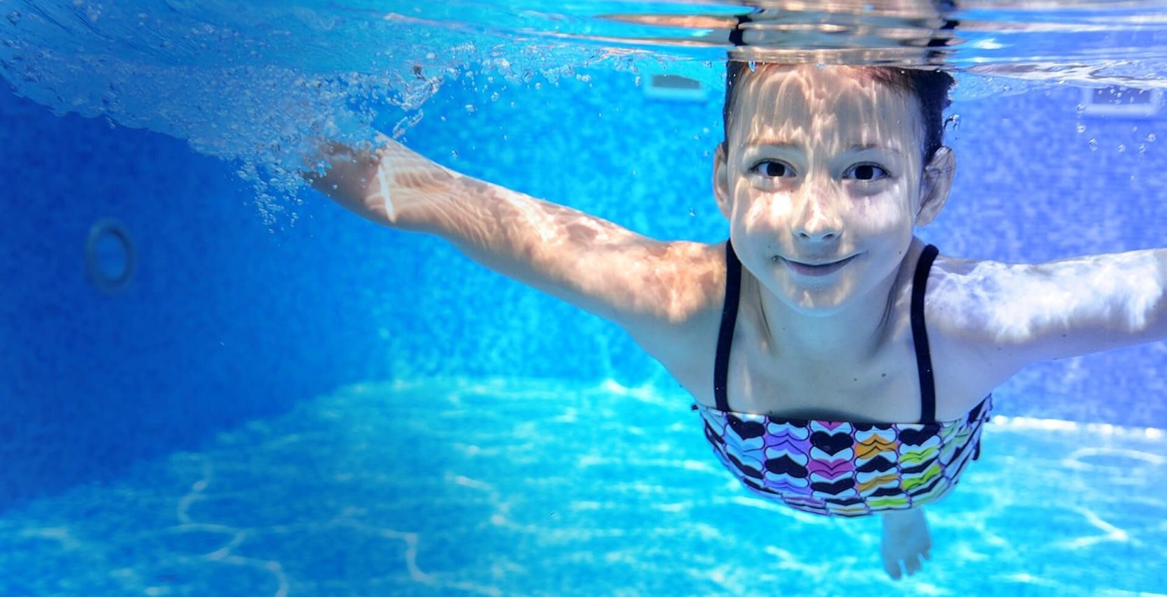 Kind schwimmt unter Wasser in einem Pool mit blauem Wasser und trägt einen bunten Badeanzug
